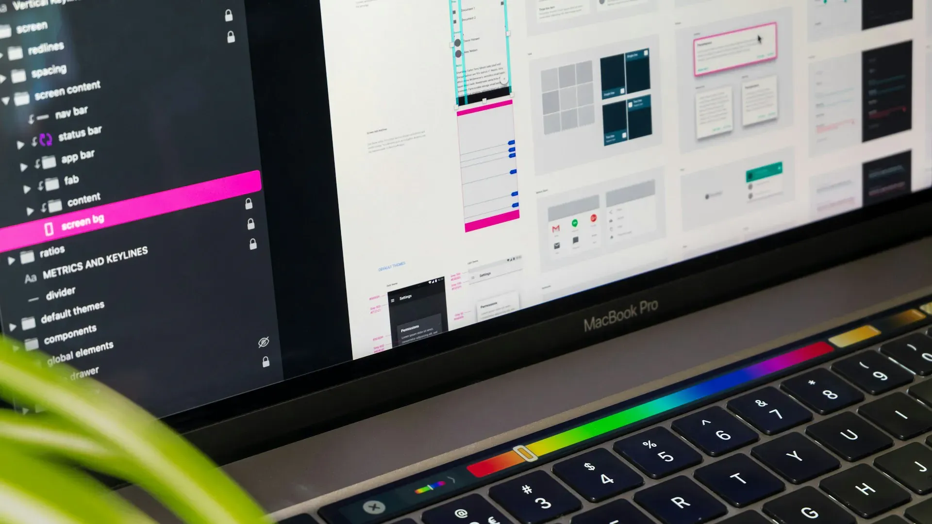 Un écran de MacBook Pro affichant une interface de conception d’UI/UX, avec plusieurs maquettes d’écrans et composants d’une application ou d’un site web. Le focus est sur un logiciel de design où l’on voit différents dossiers et éléments organisés dans la barre latérale gauche, tandis que l’écran principal montre des wireframes et des designs d’interfaces. Un coin d’une plante verte est visible en bas à gauche de l’image, ajoutant une touche de naturel à l’environnement de travail numérique.