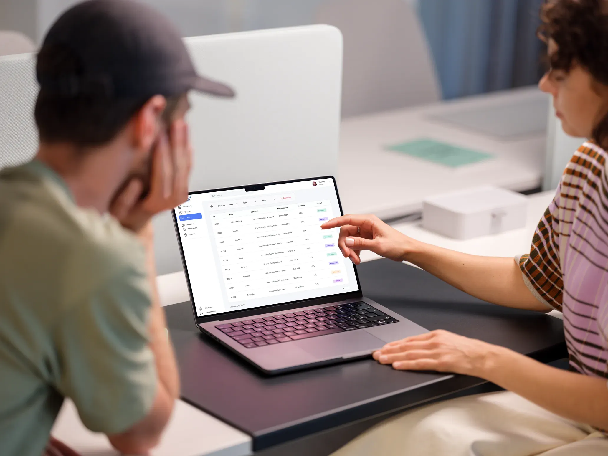 Un homme et une femme sont assis autour d’un ordinateur portable ouvert, regardant l’écran ensemble. La femme, à droite, pointe du doigt l’écran, qui affiche une interface de gestion avec des données organisées en colonnes, probablement une application de suivi ou de gestion de projet. La scène suggère une collaboration ou une discussion de travail autour des données présentées sur l’ordinateur.